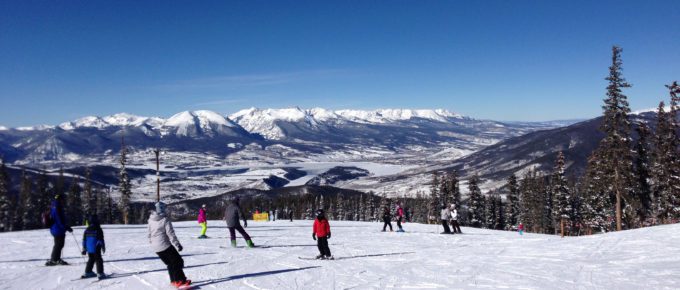 People skiing downhill in Colorado, the sky is blue and snowy mountains are in the distance.