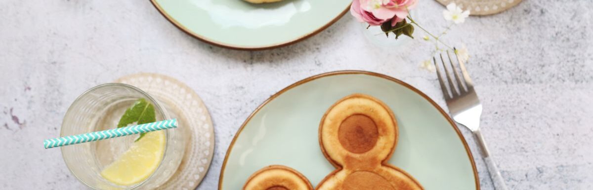 Two mickey mouse pancakes on different plates with drinks and forks on the side.