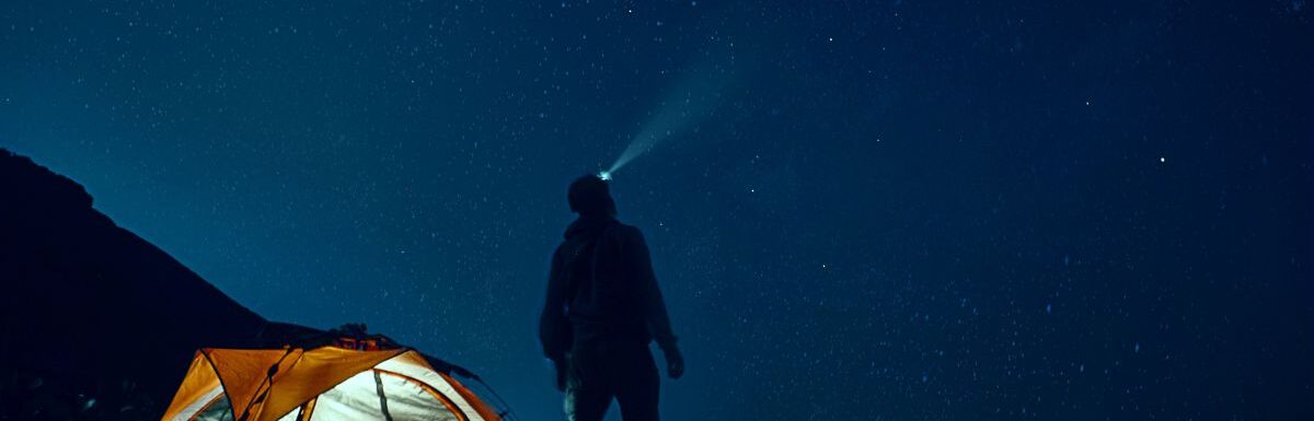 Man standing beside camping tent wearing headlamp while stargazing.
